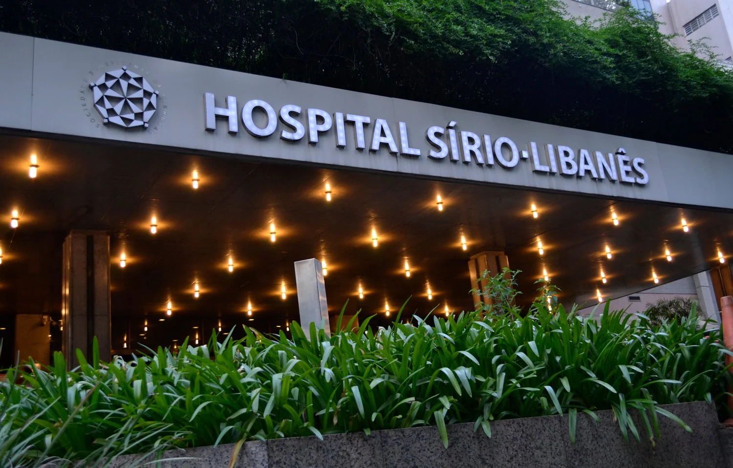 Entrada do Hospital Sírio-Libanês, com cobertura iluminada, identificação e jardim.